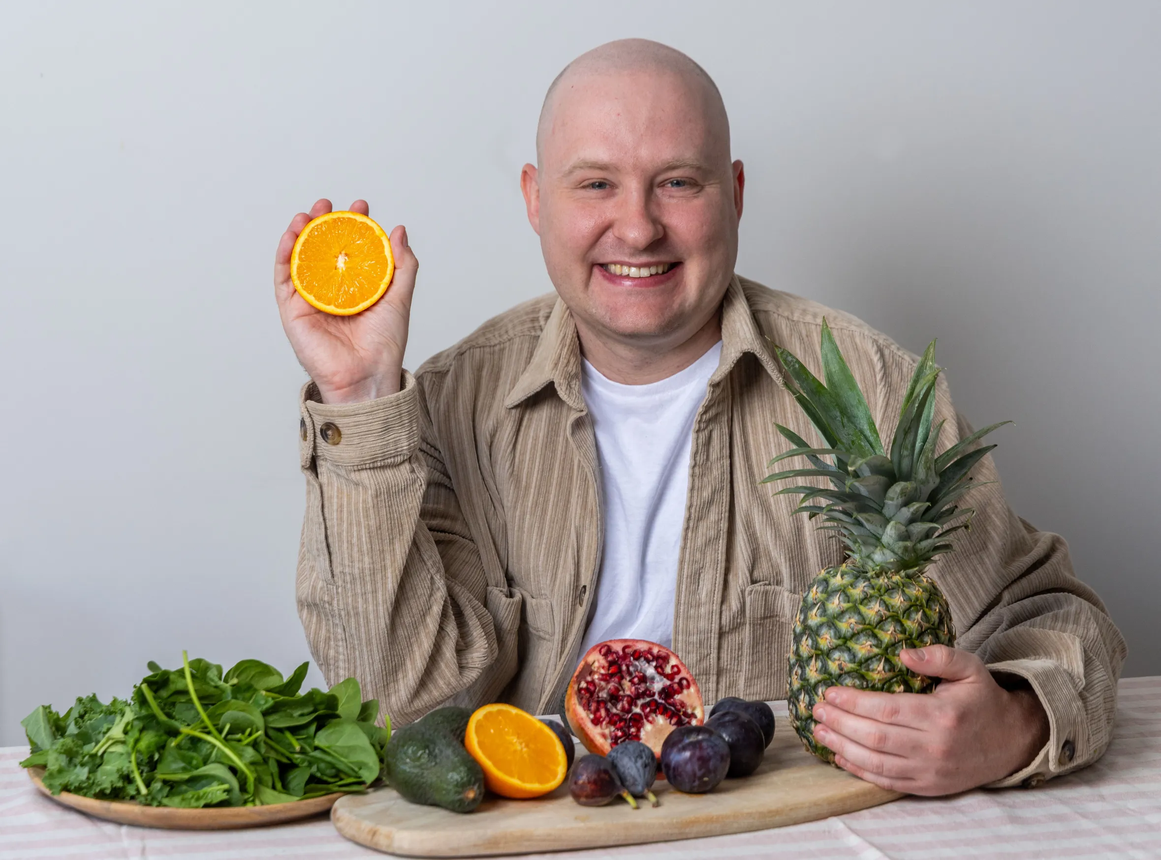Dr. David Cox smiling, holding half an orange and a pineapple, surrounded by other fruits and vegetables on a table.