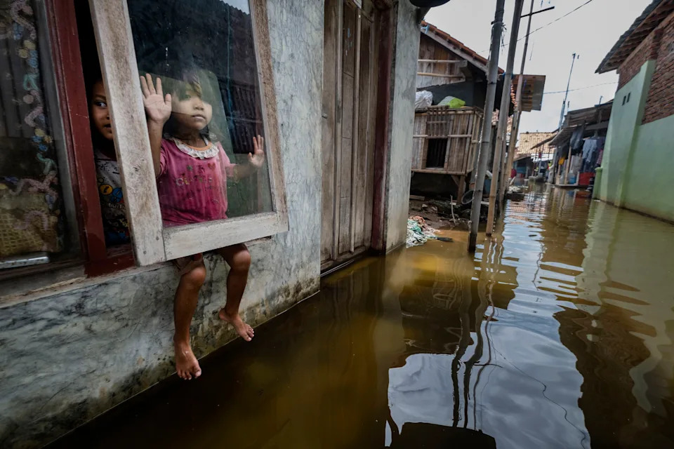 Two children look out from a window of their flooded home in Java, Indonesia, one pressing her hand against the glass and dangling her bare feet above the murky brown floodwater