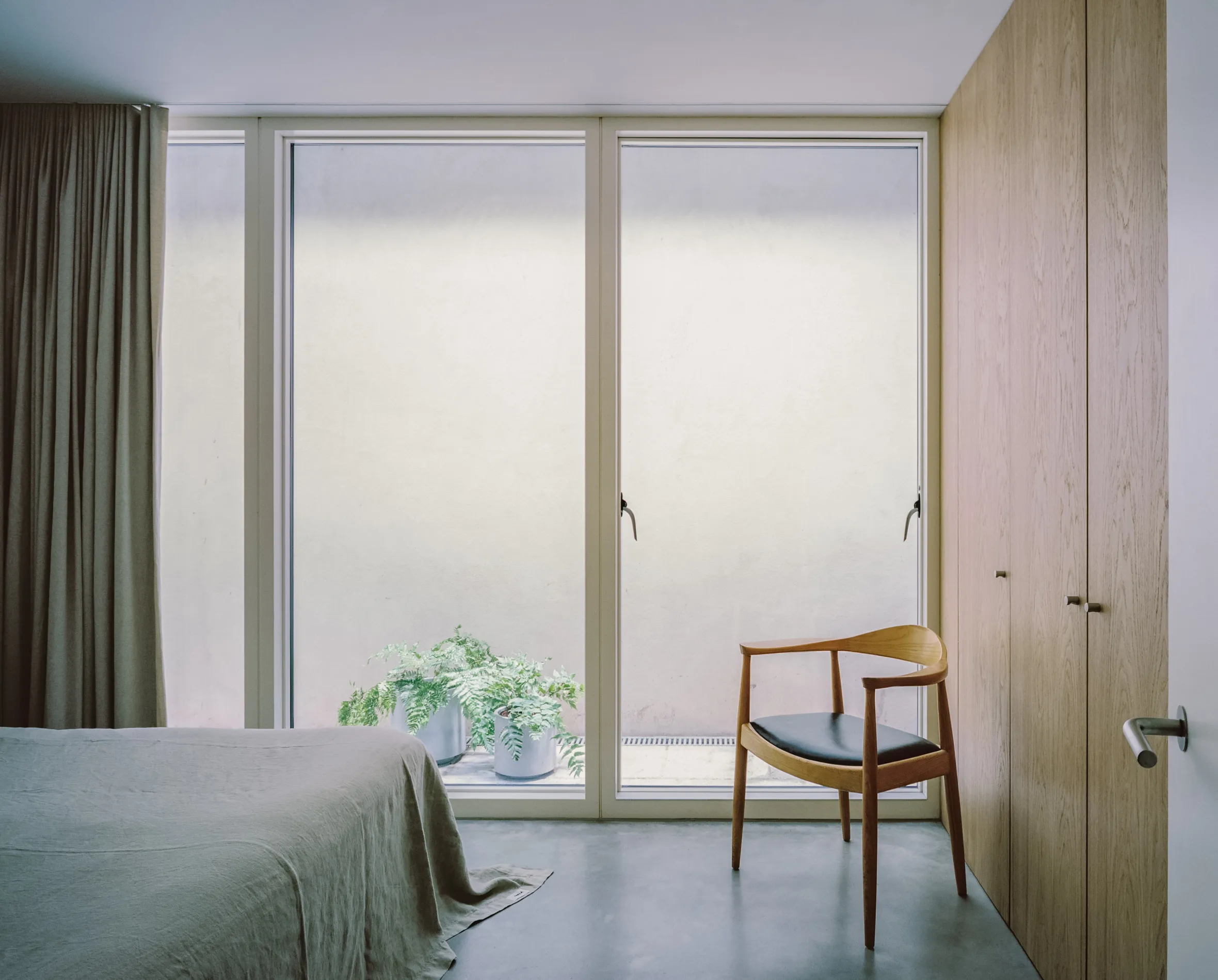 A minimal bedroom with a bed, a wooden chair, and potted ferns in front of a window.