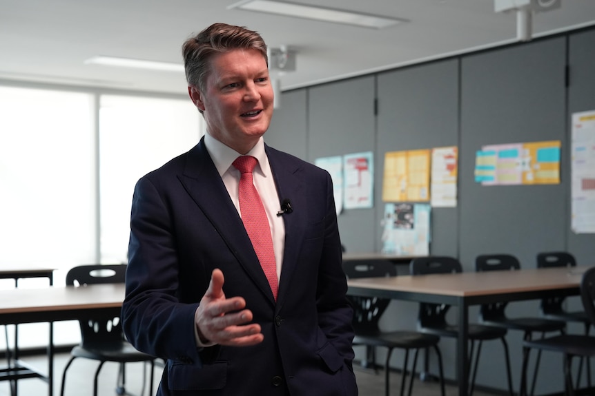 Ben Carroll talks to someone off camera, wears a dark suit, pink tie, in a classroom, desks, chairs behind.