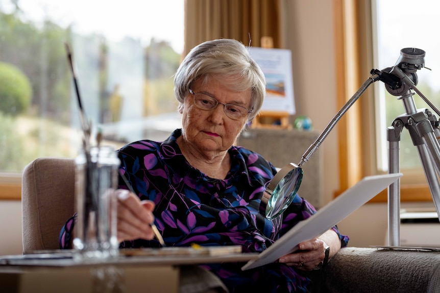 Woman sits on chair, using a magnifying glass to paint.