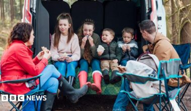 Children sitting together in the boot of a van in the woods eating packed lunch and two adults sitting on camping chairs outside.