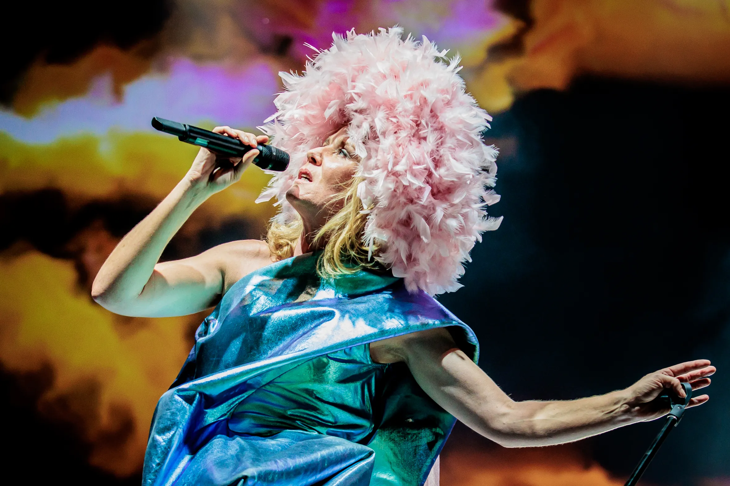 Róisín Murphy singing into a microphone, wearing a shiny blue garment and large pink feathery headpiece.