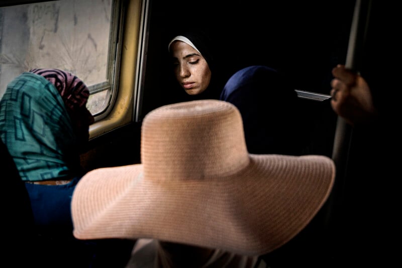 A woman in a dark headscarf sits by a window on a dimly lit bus, eyes closed. Next to her, another woman wears a patterned scarf. In the foreground, a wide-brimmed beige hat partially obscures the scene.