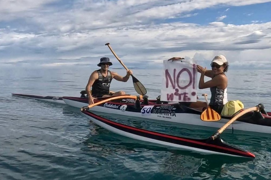 Two people sit in a canoe on open water with one woman holding a sign which says "no W2E"