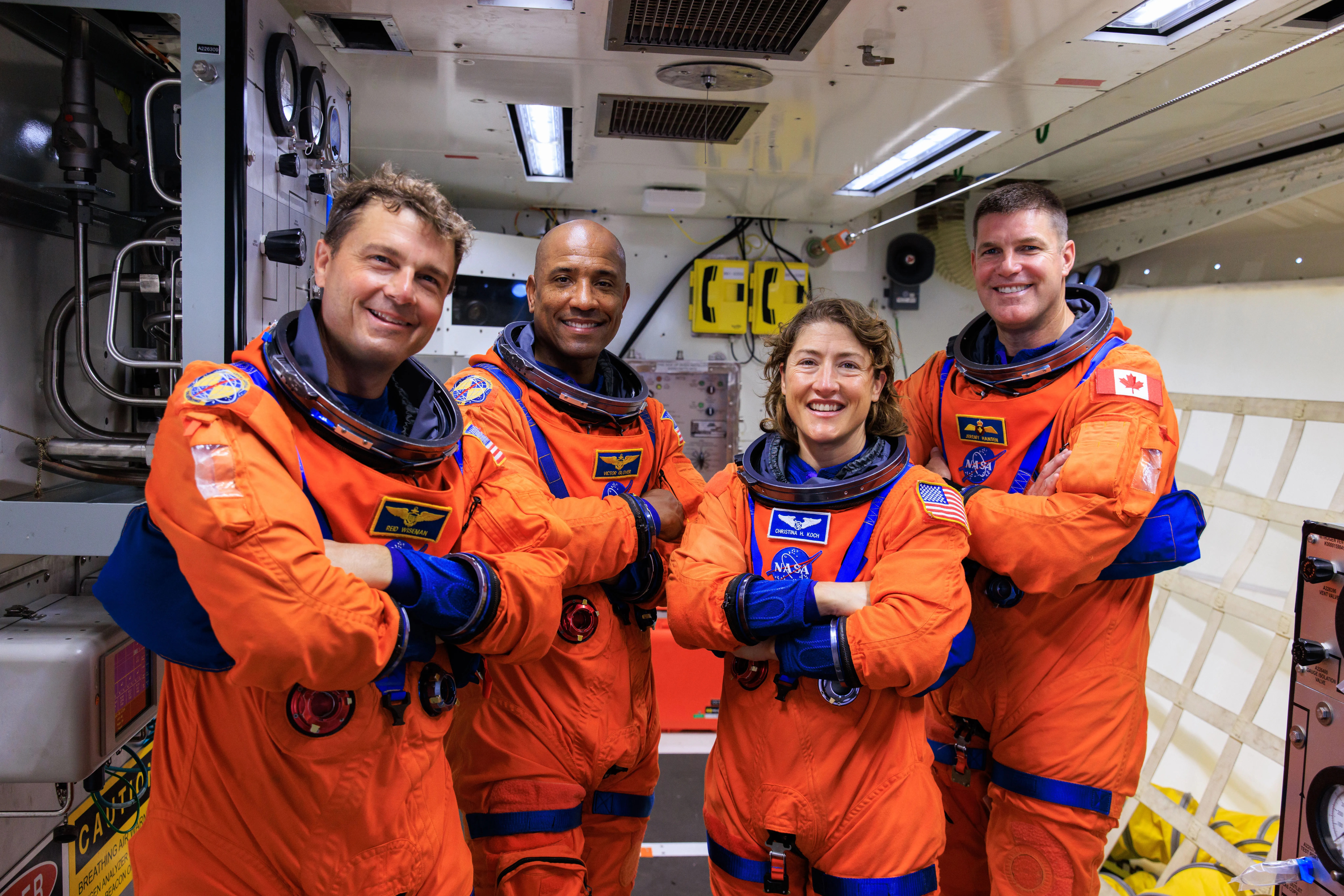 Four Artemis II astronauts in orange NASA spacesuits stand side by side inside a spacecraft module, smiling with arms crossed. Behind them, technical equipment hints at their mission to bring hope as they journey toward the moon and back to Earth.