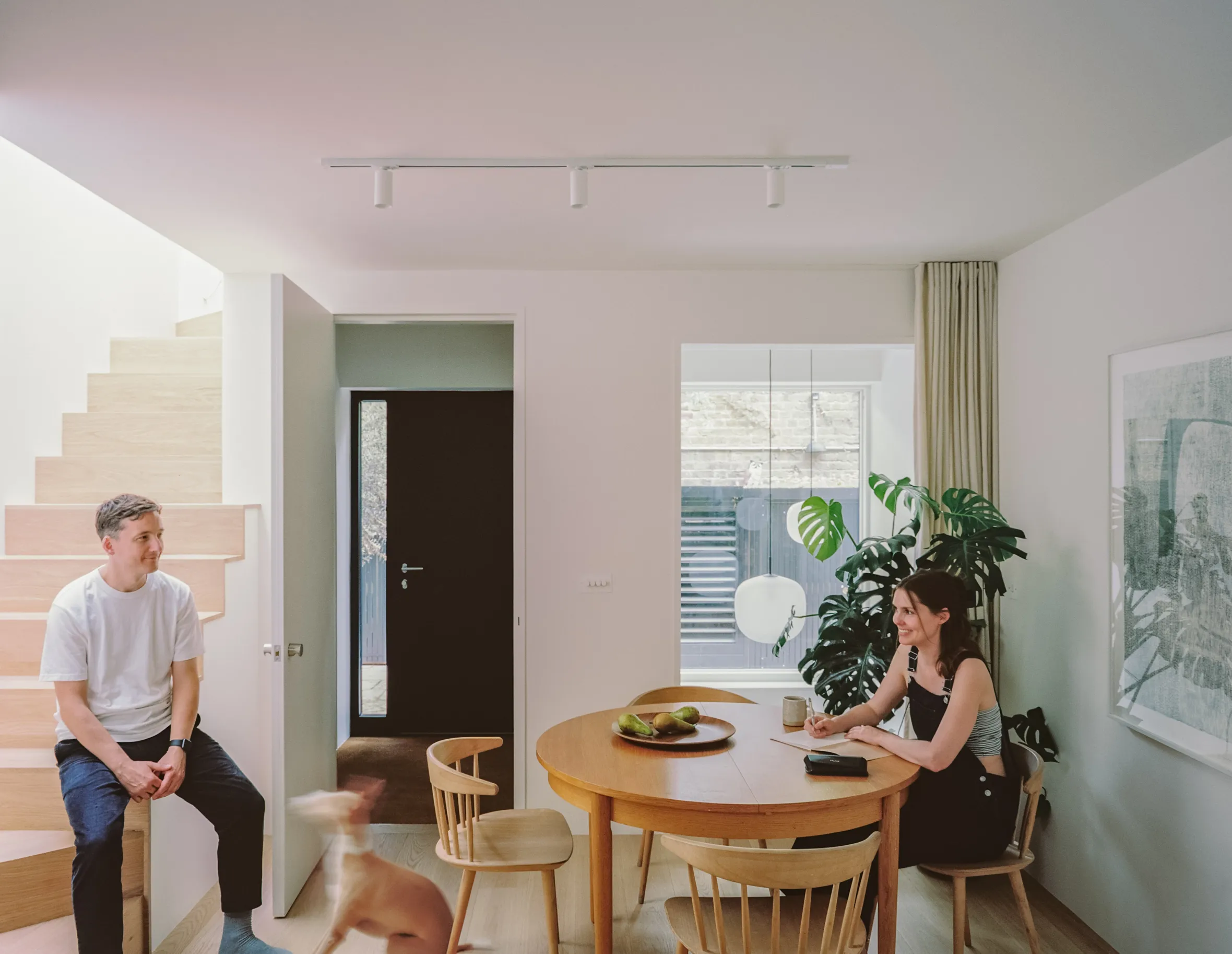 James Davies sitting on a wooden staircase and Sophie Ellis-Hayle sitting at a wooden table in their home, with a blurry image of a dog next to the stairs.