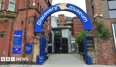 The entrance to the Discovery Museum. The venue's name is carried on an arched blue sign above opened metal gates. The building is largely built from brick but has a glass foyer.