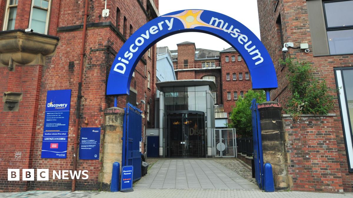The entrance to the Discovery Museum. The venue's name is carried on an arched blue sign above opened metal gates. The building is largely built from brick but has a glass foyer.