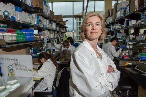 Jennifer Doudna wearing a white lab coat in a busy lab where scientist are working behind her on benches below shelves stacked tightly with boxes and supplies