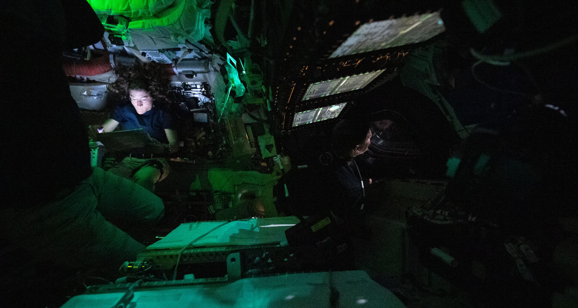 Astronaut Christina Koch is nestled into a corner of the Orion crew capsule, reading. The rest of the capsule is dark, but her face is lit by the glow from her tablet. Two of her other crew members can dimly be seen looking out the window and floating in the foreground.