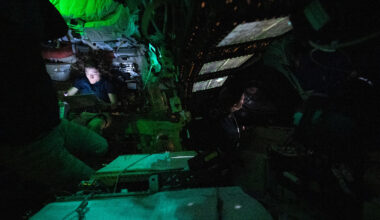 Astronaut Christina Koch is nestled into a corner of the Orion crew capsule, reading. The rest of the capsule is dark, but her face is lit by the glow from her tablet. Two of her other crew members can dimly be seen looking out the window and floating in the foreground.