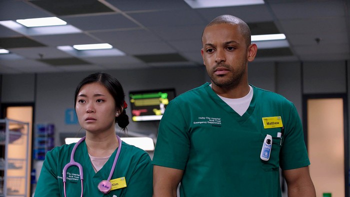 553524,TITLE:Casualty Jasmine Bayes as Kim and Aron Julius as Matty stood next to each other in green scrubs.