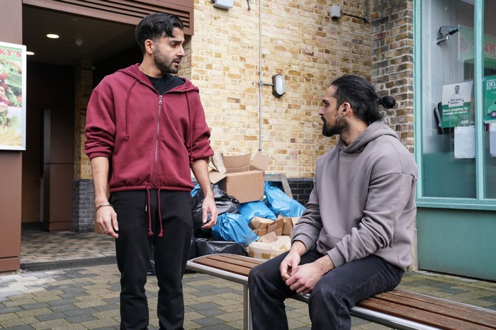 Vinny stands over Ravi beside some boxes and bin bags in a scene from EastEnders
