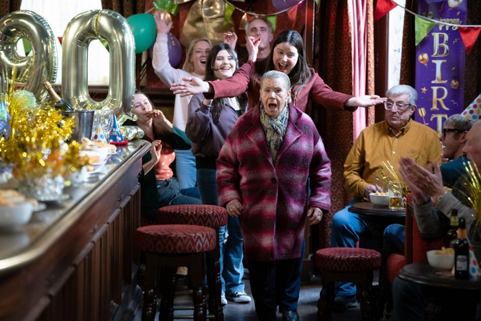 557194,TITLE:Eastenders Mo walks into The Vic surrounded by friends and family, with a shocked expression on her face and some big balloons with the number '90' on them in a scene from EastEnders