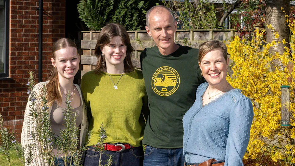 A lady in a blue blouse, from the earlier picture. She stands with a tall bald man with a dark green t shirt. Two girls who appear in their teens or early 20s stand next to them. They all have arms around each other and are smiling at the camera.