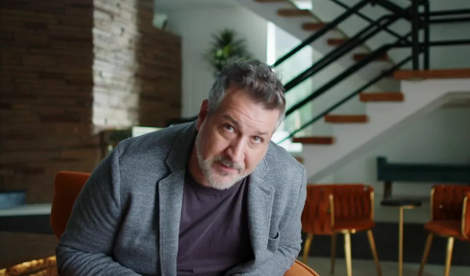 A man with short hair and a beard is sitting in a modern interior wearing a casual blazer and shirt, looking toward the camera