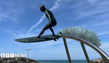 A statue of a surfer on a surf board suspended on a steel pole with the sea behind it. Behind the surfer is a bronze wave symbol