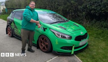 A man with brown hair and glasses and wearing a green hoodie, dark green trousers with black boots is stood in front of his bright green car. He has his thumbs up and is looking straight at the camera.
