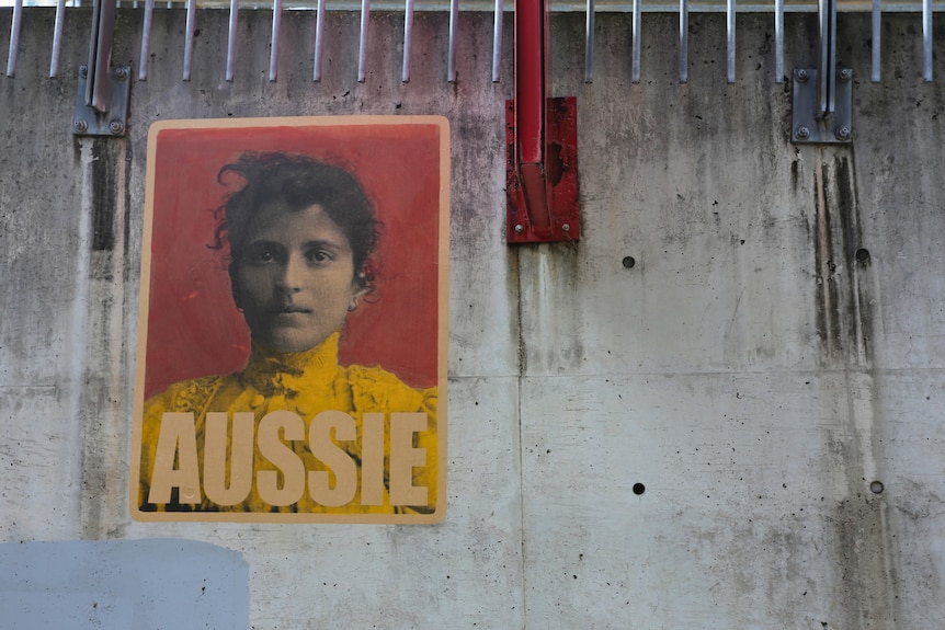 A poster of a young woman with the word Aussie at the bottom, stuck on a grey wall.