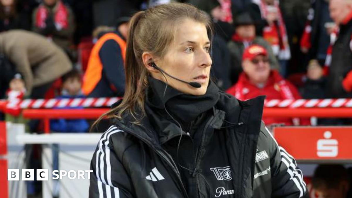 Marie-Louise Eta, wearing a black coat and top with a black microphone and headset in her right ear, on the touchline with fans in red and dark clothing in the background