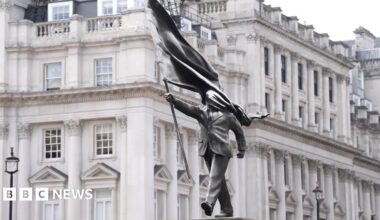 A grey statue of a man wearing a suit, marching forward, carrying a flag that covers his face