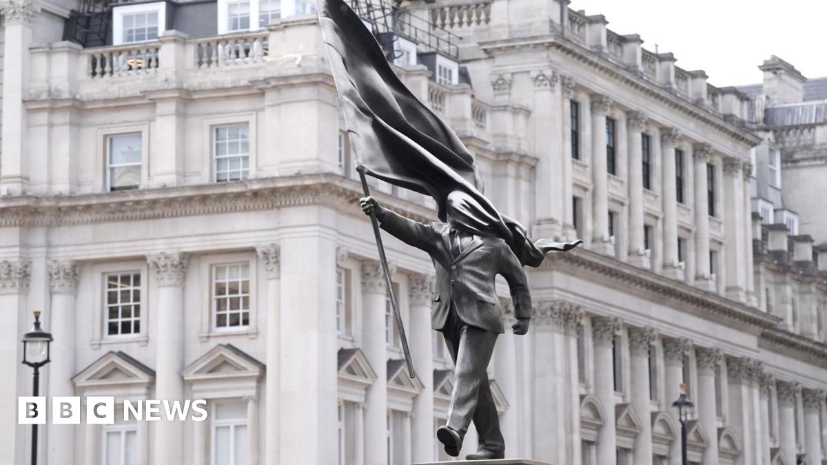 A grey statue of a man wearing a suit, marching forward, carrying a flag that covers his face