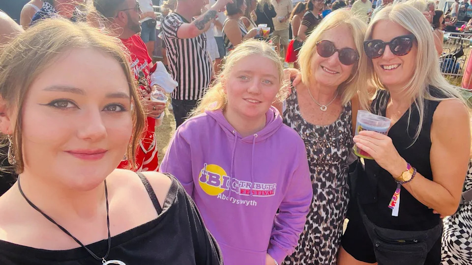 Helen smiling before she became unwell with her daughter and two granddaughters, wearing sunglasses and a leopard print top. They are stood at a festival in the sun, with people in the background. 