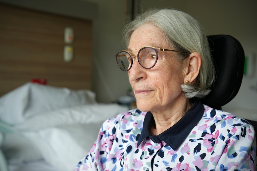 A woman with grey hair and glasses sits in a wheelchair in a hospital room.