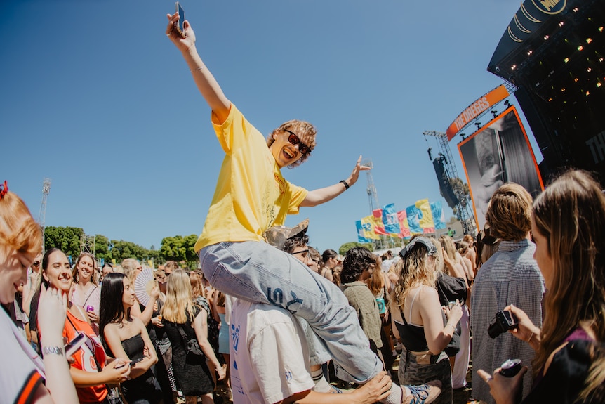 A man on the shoulders of someone else at a music festival with a crowd of people around him