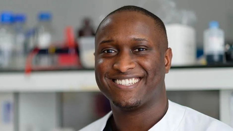 A man with a shaved head, short goatee and white shirt, in front of a blurred lab background, smiles at the camera.