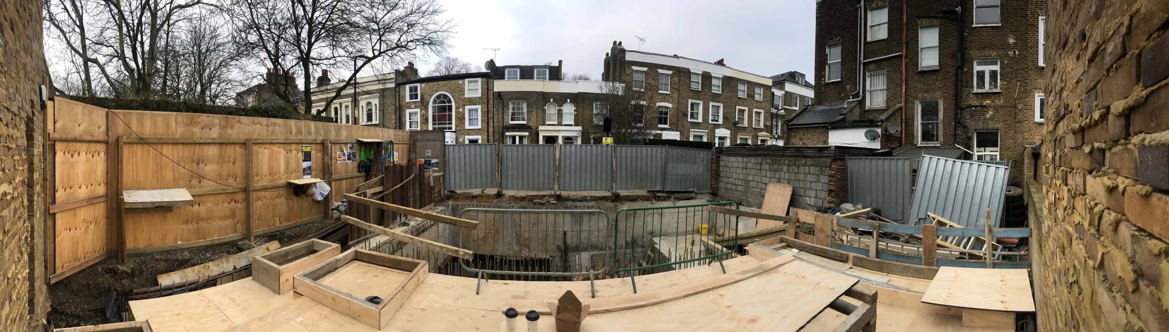 A panoramic view of a construction site for Greenview House with excavation work and plywood forms in the foreground, and residential buildings in the background.