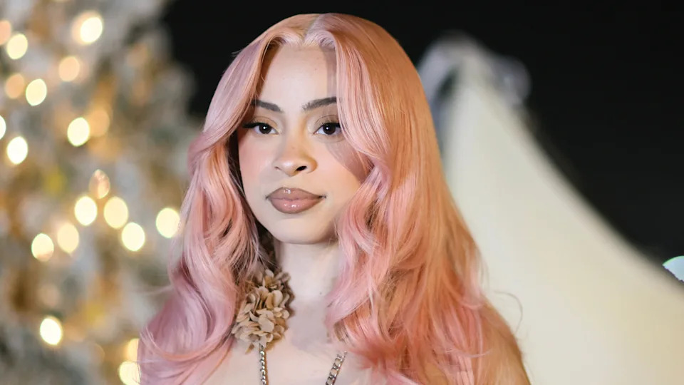 Ice Spice with long, wavy pink hair, wearing a floral necklace, stands in front of a blurred background with lights.