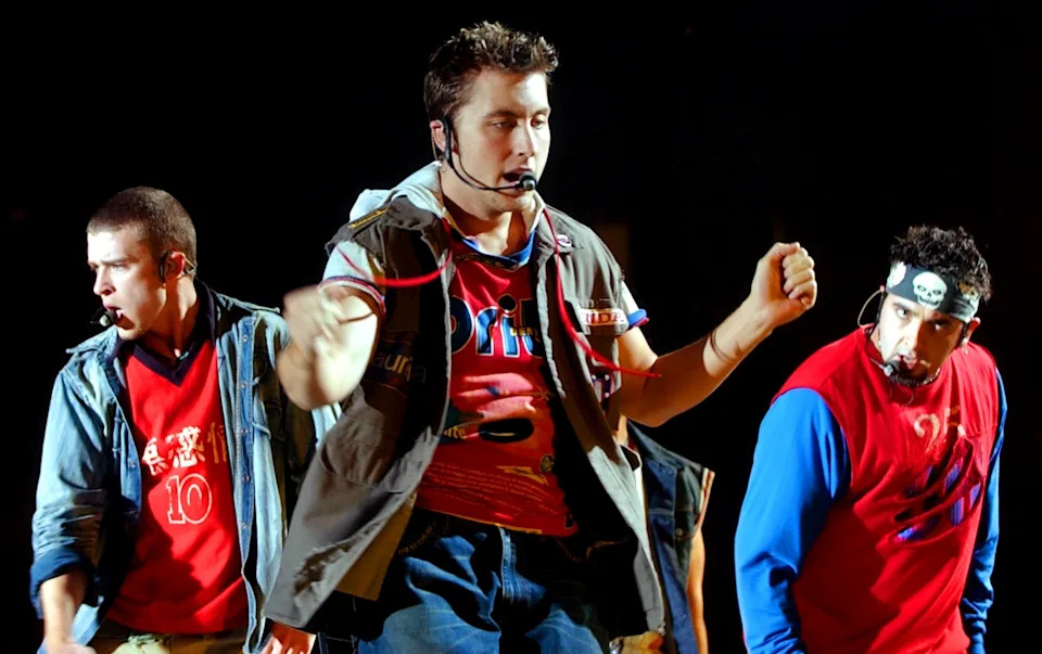 Three performers on stage, wearing casual layered outfits, each with a microphone headset, actively dancing during a concert
