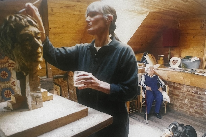 The artist with long grey hair in her studio working on a bust of an older woman who is seated behind her