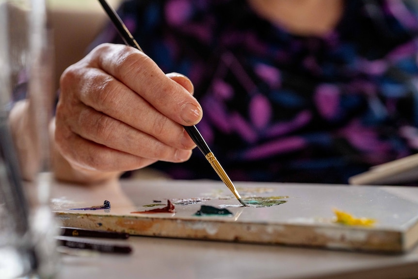 Woman sits on chair, using a magnifying glass to paint.