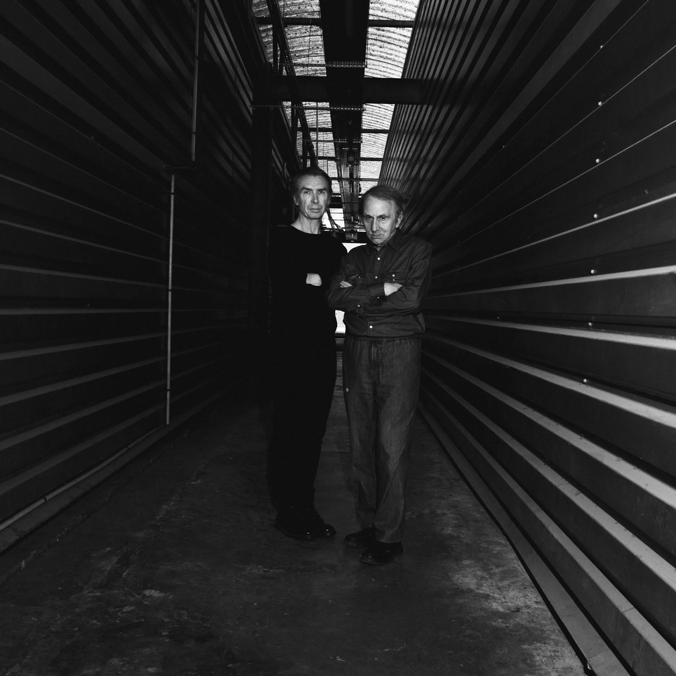 Black and white photo of Michel Houellebecq and Freederic Lo standing with arms crossed in a narrow, industrial-looking hallway.