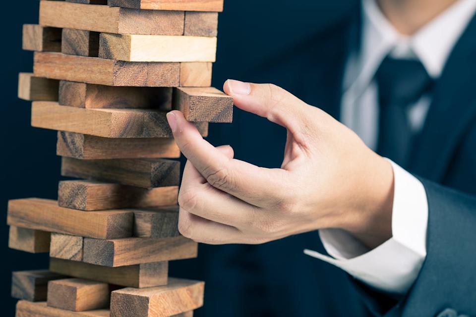 A person in business attire pulls a wooden block from a wobbly tower, illustrating a shaky investment thesis in a stock. 