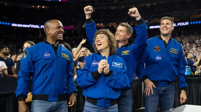 A foto mostra quatro astronautas da missão lunar Artemis 2 da Nasa em pé em uma arena esportiva coberta e movimentada, vestindo jaquetas de voo azuis brilhantes com emblemas e logotipos da missão. Eles estão em primeiro plano, sorrindo e comemorando, com uma mulher no centro juntando as mãos e dois dos homens erguendo os punhos no ar em sinal de celebração. A multidão atrás deles está ligeiramente desfocada, mas preenche as arquibancadas, sugerindo um grande evento esportivo. 