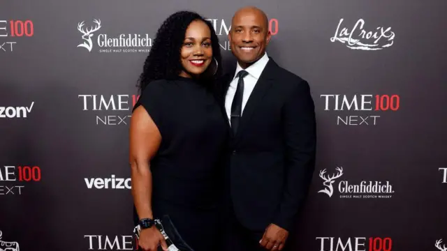 Victor Glover está à direita, de terno escuro, camisa branca e gravata preta fina, com o braço delicadamente em volta das costas de Dionna enquanto posam juntos no tapete vermelho. Dionna, à esquerda, usa um vestido preto simples na altura do joelho e carrega uma bolsa de mão estampada na mão direita. Eles estão próximos, de frente para a câmera, contra um fundo escuro com a inscrição “TIME100 Next” e logotipos de patrocinadores, criando uma atmosfera formal, porém descontraída e festiva.