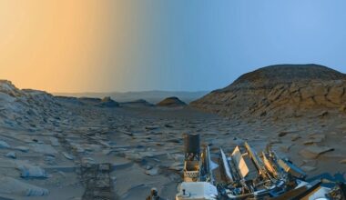 Ancient lake on Mars: Rocky terrain with a mesa in the distance on the right and a yellowish and bluish sky. Part of a mechanical rover is also on the right in the foreground.