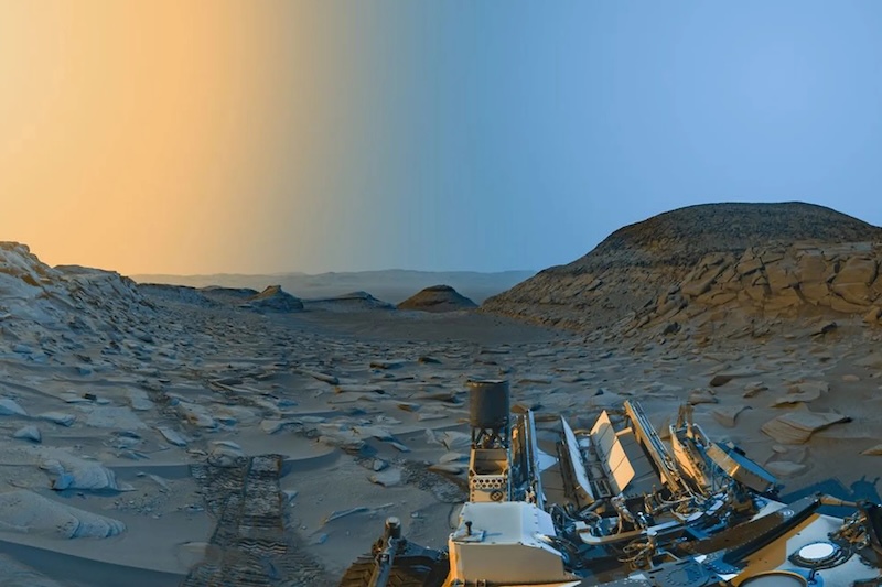 Ancient lake on Mars: Rocky terrain with a mesa in the distance on the right and a yellowish and bluish sky. Part of a mechanical rover is also on the right in the foreground.