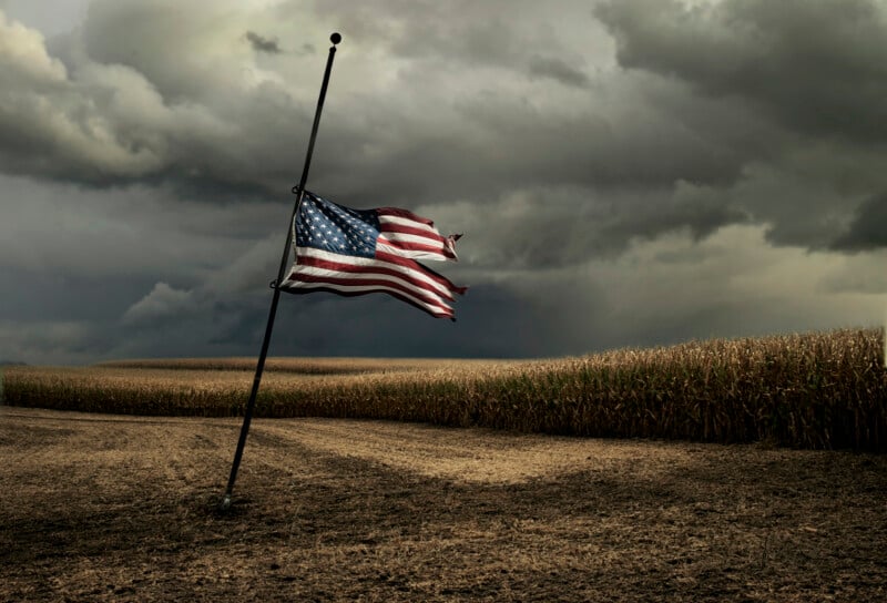An American flag at half-staff waves in the wind on a barren field beside a cornfield under a dark, cloudy sky, creating a somber and dramatic atmosphere.