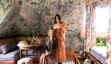 Two women pose in a cozy, patterned room with floral wallpaper, a wooden table set with flowers, and colorful curtains framing a bright window.