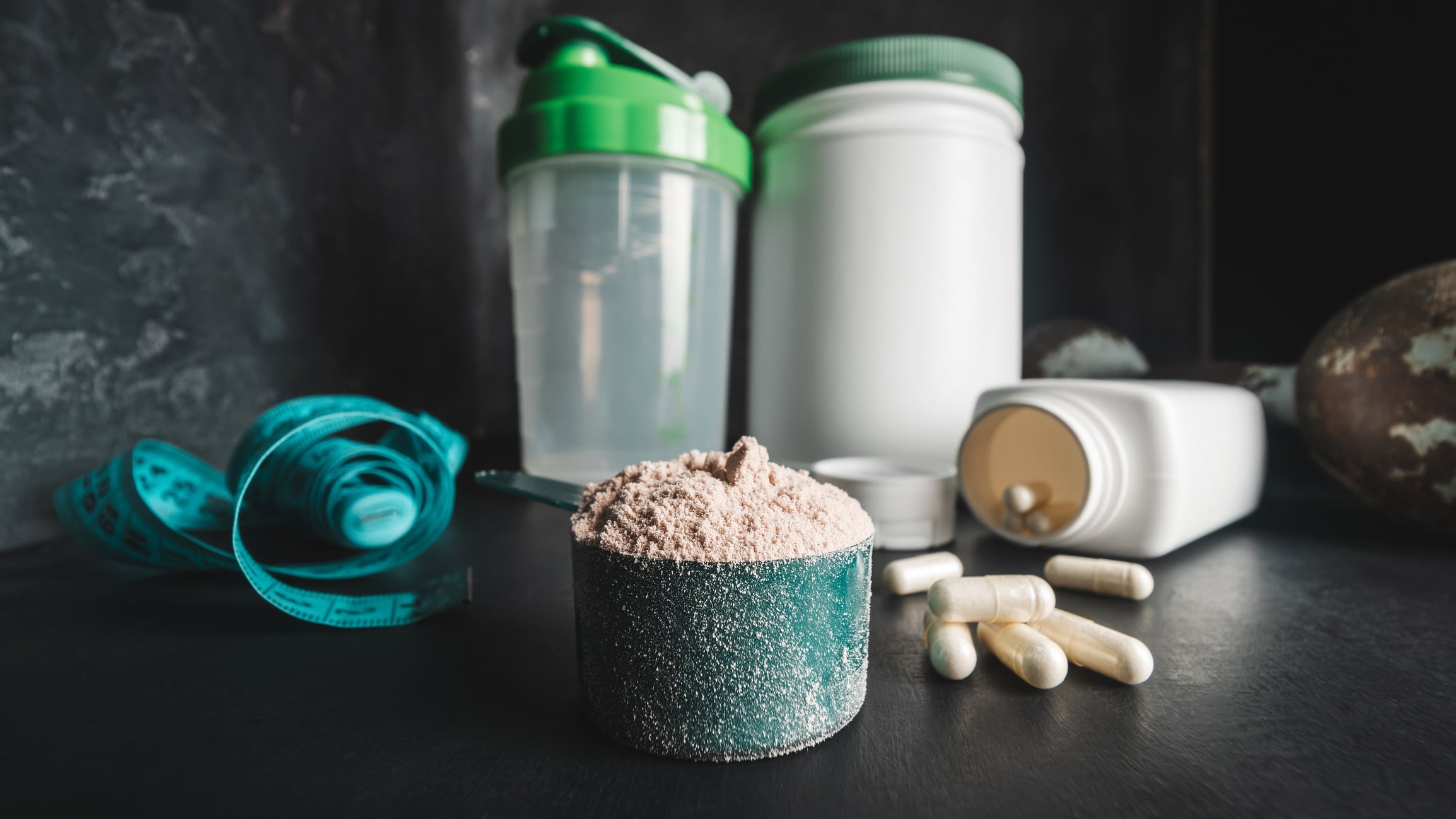 Chocolate whey protein powder in measuring spoon, white capsules of amino acids and vitamins, measuring tape, old rusty dumbbell, plastic shaker on dark background. bodybuilding food supplements.
