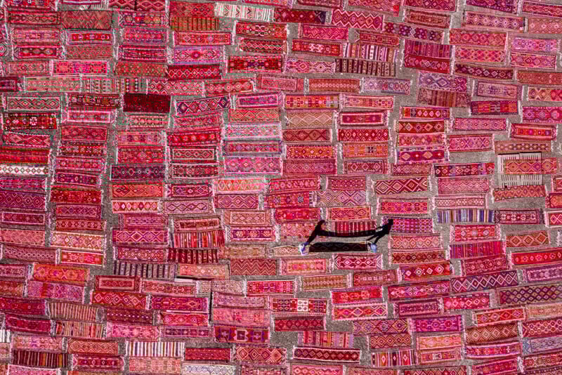 A large display of vibrant, patterned red and pink rugs covers a wall, with a single black cushioned seat positioned near the center. Each rug features unique traditional designs.