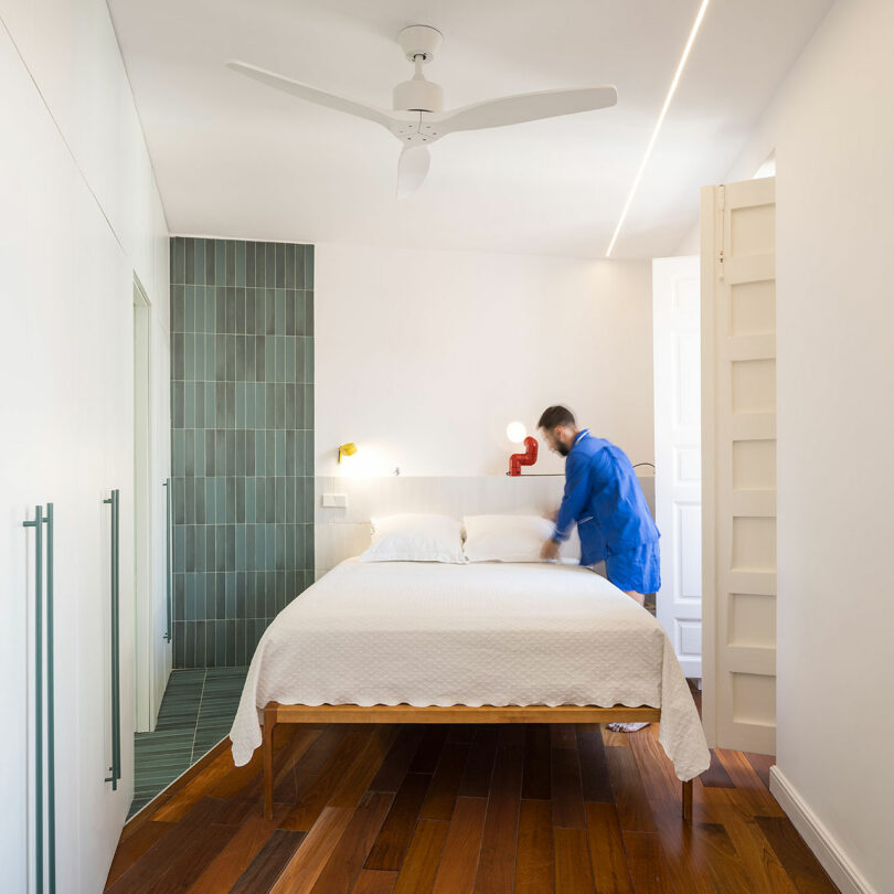 Person in a blue outfit makes a bed in a minimal, modern bedroom with white walls, green tiled accent, wooden floor, and a ceiling fan.