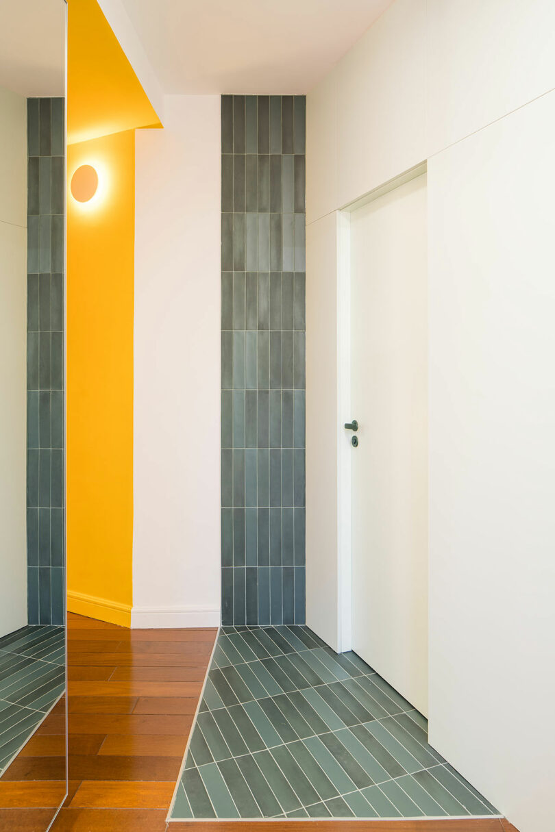 A modern hallway with a white door, vertical gray tiles on the wall and floor, a yellow accent wall, and wooden flooring. A mirror reflects part of the space.
