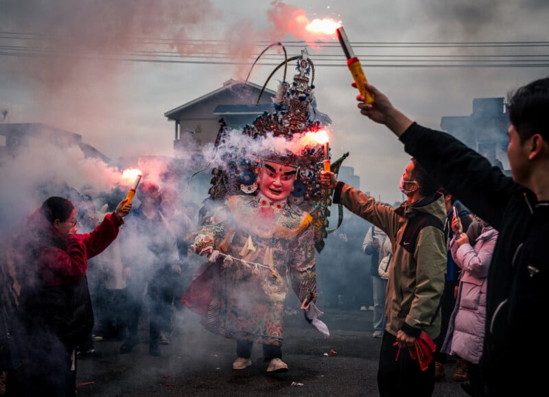 A person in elaborate traditional costume and mask stands amid dense smoke, while people around them hold lit flares during an outdoor celebration or festival. Houses and power lines are visible in the background.