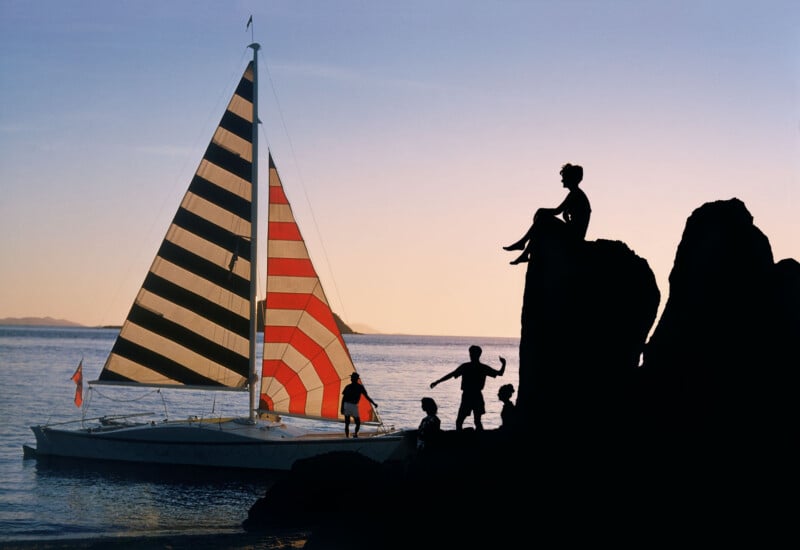 A sailboat with a striped black, white, and red sail is anchored near the shore at sunset. Four people are silhouetted on nearby rocks, one seated above the others, with the calm water in the background.